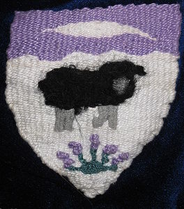 Philippa Llewelyn Schuyler Argent, a sheep statant guardant contourny sable, in base a clump of lavender vert flowered purpure, on a chief invected purpure a quill pen reversed argent. (registered 1/93). Maker: Philippa Llewelyn Schuyler. Notes: The shield is tapestry-woven with pearl cotton. The lavender is worked with French knots.