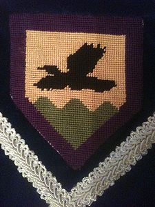 Raven of Heronsmarsh Or, a raven volant wings addorsed sable and a base wavy vert, all within a bordure purpure. (registered 7/1996). Maker: Raven of Heronsmarsh.